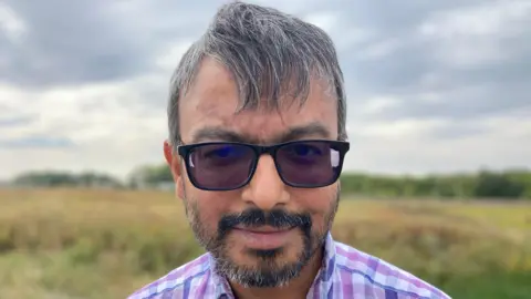  Helen Mulroy/BBC Saqhib Ali, wearing sunglasses, and a checked shirt, in a field in Wixam's, Bedfordshire