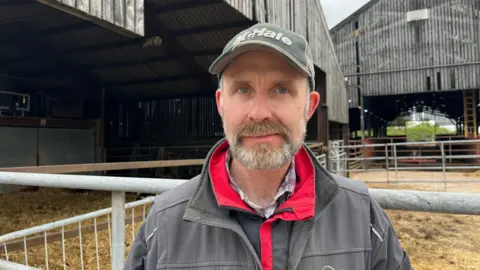 Ellen Knight/BBC Photograph of Chatham looking into the camera with a neutral expression. He has a short, dark grey beard. He is wearing a red and black waterproof jacket with a grey overall over the top. Chatham is also wearing a dark green cap with 'McHale' written on it in white lettering. He is stood in front of two huge barns, clad in corrugated iron. There are metal gates and fences in front of the entrances, with straw strewn across the floors. 