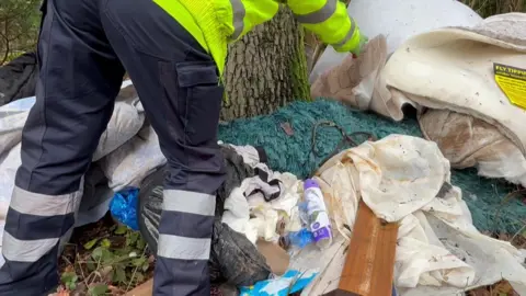 A council worker sorting through fly-tipped rubbish. He is wearing a fluorescent green top and blue trousers with white bands. He is photographed from the waist down.