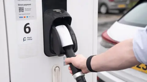 A hand reaches for an electric charging point, which is white, with the number six on it, there is a white car in the background.