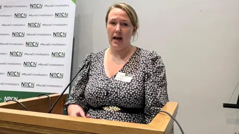 BBC/RICHARD EDWARDS Robyn Munt, vice president of the National Farmers' Union, who told today's rural crime cconference about the "human cost" of wildlife crime, is pictured at the conference speaking podium. She is wearing a black dress with a white floral print and has blonde hair which is tied back into a ponytail