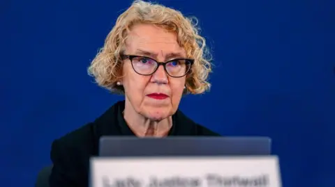 PA Media Lady Justice Thirlwall has blonde bobbed hair and is wearing black framed glasses and a dark coloured suit. She is seated in front of a laptop, with a sign in front of her saying her name. There is a blue background.