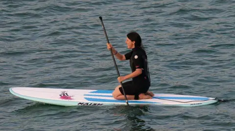 Getty Images A woman kneeling on a paddleboard at sea, She is wearing a black shorty wetsuit. The board is white, blue and green.
