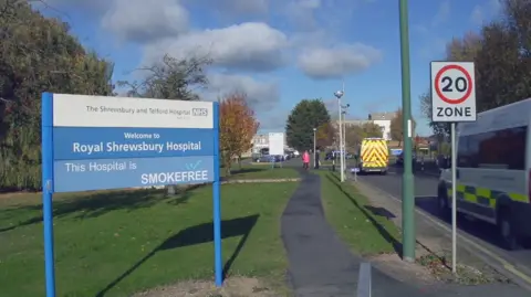 BBC A blue and white sign for the Royal Shrewsbury Hospital. To the right of it is a road, with two ambulances driving along it, towards the hospital building