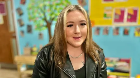 Martin Giles/BBC A woman is sitting in a room for children. It has a bright coloured wall painted blue. There are pictures, photos and tree painted on it. Chelsea is wearing a white top and matching jacket. She is smiling at the camera. 