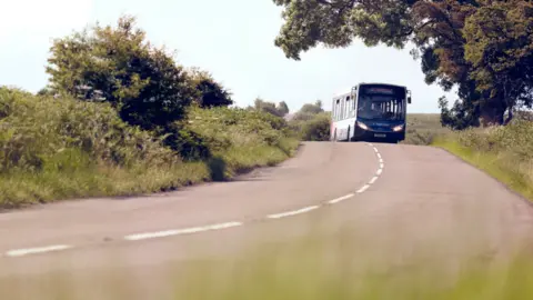 A single-deck bus travels along a country road. Green bushes are on either side of the road. 