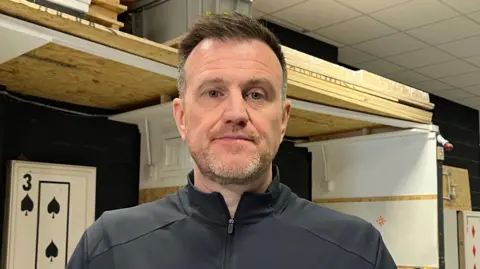 A man wearing a black jacket is standing in front of stacks of wood, including a piece that is designed to look like a playing card.