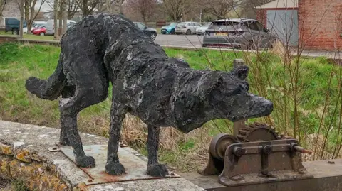 The black cast dog sculpture standing on top off a wall with the weir mechanics in the background. The dog resembles a border collie when it stalks sheep. Its head is low, tail is down and ears are forward.