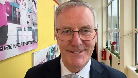 Mike Nesbitt is wearing square glasses, he has a dark navy blazer on, a white shirt and a grey tie. Behind him can be seen a yellow walled hallway with Christmas decorations. 