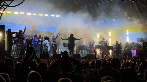 The stage at Summer Fest at the Beach in Weston-super-Mare. There are musicians on stage and a large crowd can be seen watching them.