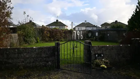  general view of the former site of the Bon Secours Mother and Baby home and the memorial garden. There is a gate and stone wall with a garden and the number '796' fastened to the far wall.