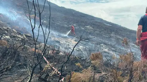 Derbyshire Fire and Rescue Service Burnt moorland with a firefighter spraying water on the ground.