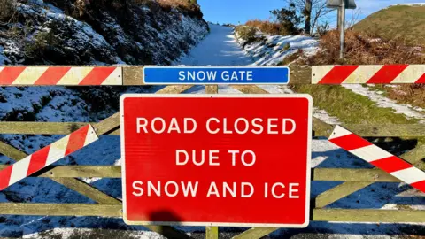 Snapper Simon / BBC Weather Watchers A closed gate with a sign on saying: Snow Gate. Road closed due to snow and ice . Beyond it is a narrow snow-covered track climbing up a hill