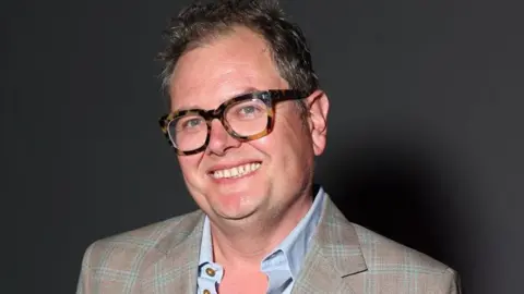 Alan Carr in a brown and light green checked suit jacket with a blue shirt underneath and brown, thick-rimmed glasses.
