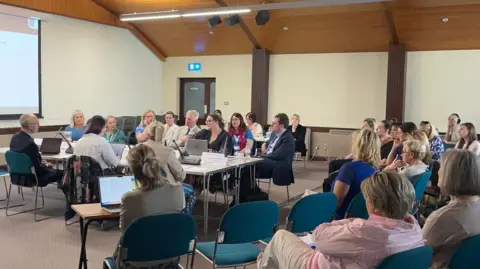Politicians sit around a table in a town-hall type setting. Members of the public look on. Tom Binet is sat centrally. 