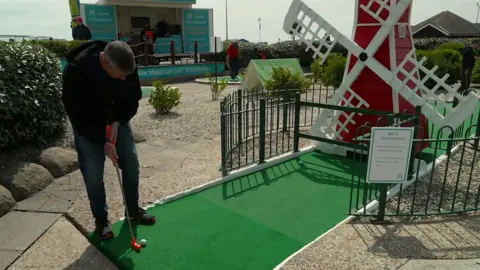 A man holding a golf club preparing to hit a ball on a crazy golf hole. The course features a large red and white windmill.