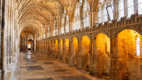 Gloucester Cathedral The cloister at Gloucester Cathedral. It is a hallway made of intricate stone arches and the windows on the right are lit with orange lighting.