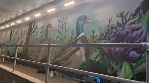 Duck, purple flowers and green plants painted on the side wall of an underpass. There is a small wall with metal railings on top in front of it. 