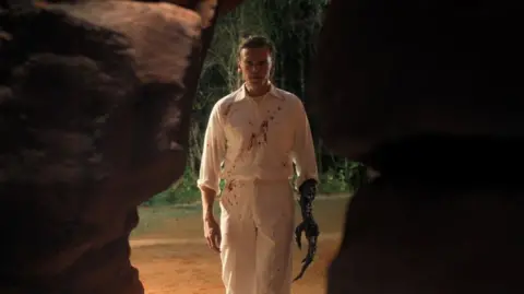 Netflix Henry Creel in a white, bloodstained shirt and trousers, with Vecna's arm starting to grow - it is burned and has long, sharp fingers and claws