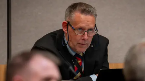 Senedd Cymru Gaz Thomas wearing glasses, a multi-coloured tie, a blue shirt, and a black suit.