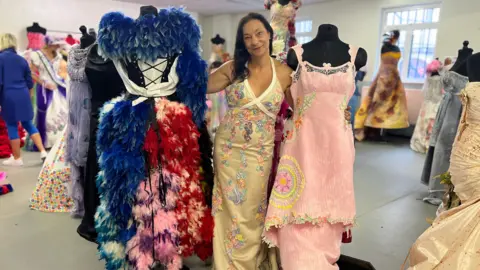 Tany's sweater next to two dresses - one is a blue, red and pink dress covered in feathers. The other is a pink dress with some flowers and a yellow circle decorating it.