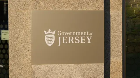 A gold Government of Jersey sign on the outside of its main headquarters. The sign is on a stone wall with an automatic door on the left hand side.