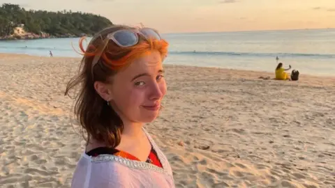 Luke Walsh A young woman stands on a beach. She is smiling at the camera.