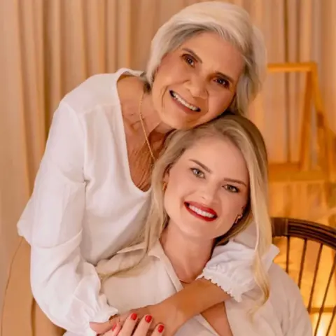 Fekete family archive A young woman with long blonde hair sits on a chair. An older woman with chin-length grey hair stands behind her, with her arms around the seated woman's shoulders. They are both wearing white tops and are smiling. 