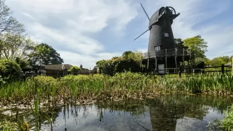 BBC Weather Watcher dieterswanderings Bursledon Windmill - a black windmill with a large pond stretching beneath full or reeds.