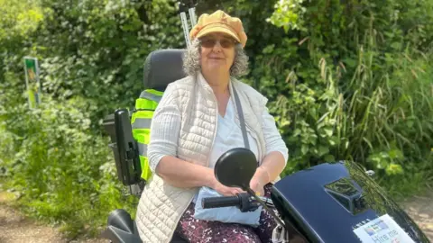 A woman with grey hair, wearing a yellow hat and stone gilet, sits on a mobility scooter. She's smiling, and behind her are crutches. She is outside, on a path lined with green bushes