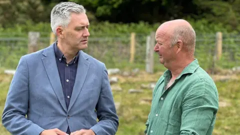 John Renner and David Smith are facing each other and talking in a field. Mr Renner is wearing a green shirt and has balding, grey hair. Mr Smith has short grey hair and is wearing a light blue blazer and dark blue, open-necked shirt