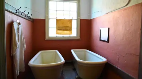 A narrow historic bathroom with two white bathtubs side by side, reddish‑brown lower walls, and a large window at the back. A long white garment hangs from wall hooks, and a yellow cloth with printed text is pegged across the window.