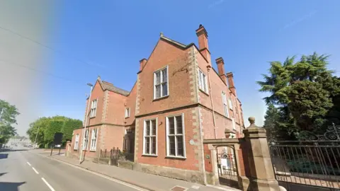 Bass House, a red brick building with ornate railing alongside it, fronts onto a main road. Behind the railings is a tree and in the distance to the left of the image there is a road junction.