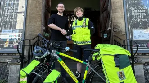A bike stands outside a church. It says AMBULANCE SERVICE on the bike and has hi-vis bags at the front and back of it. There are two people standing behind it shaking hands. A man to the left has dark hair, a beard, glasses and a dark top and jeans. To the right is a person with short fair hair, dressed in an ambulance service hi-vis tabard and black top and shorts. Both are smiling.