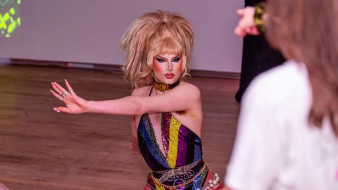 The drag queen UGLY wearing a multi-coloured sequined leotard doing an exercise move