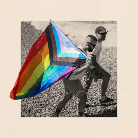 BBC A black and white photograph of two people walking outside, one of the men is holding a pride flag, with another man walking next to him in a baseball cap 