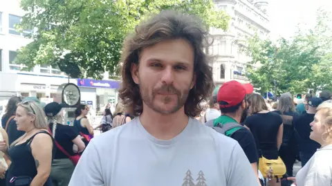 A man, with shoulder length brown hair and brown stubble, looking at the camera. He is wearing a light blue t-shirt. There is a crowd of people behind him.
