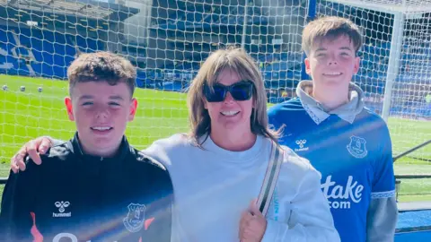 Family handout group photo of Rita flanked by her two sons in Everton jerseys, in front of the pitch at Goodison Park.