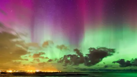 The night-sky is illuminated in hues of green, pink and purple above the clouds, as part of the northern lights. A coastline and the ocean line the bottom of the frame, with bright yellow orbs of light illuminated at the coast.