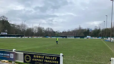 A football pitch, with advertising hoardings around it. There are two green stands, one at the far end behind the goal, and one on the left of the pitch. A man in high vis walks on the pitch at the corner of the penalty area