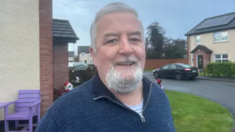 A man looking at the camera, he has white hair and beard. He is wearing a navy quarter zip
