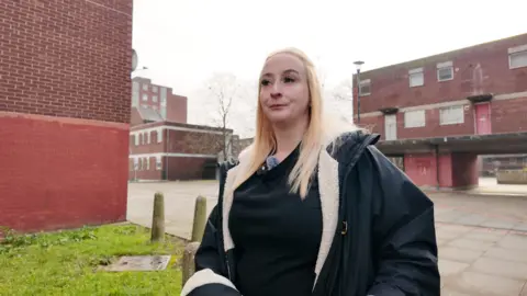 Danielle, a blonde woman in a winter coat, looks wistfully away from the camera against the backdrop of a red-brick housing estate