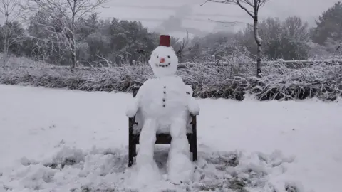 Dartmoor Charlie A snowman is sitting on a wooden chair in a  snow-covered garden. He is wearing a hat and has a carrot for a nose and stones for his eyes, mouth and buttons down his stomach.