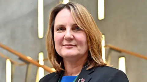 Michelle Ballantyne, with light brown highlighted hair, looks at the camera in a medium-close-up. She in wearing a dark jacket and blue top and is in the Scottish Parliament building. 