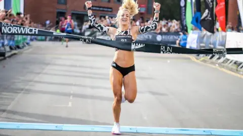 Norwich City Community Sports Foundation/Epic Action Imagery Holly Archer, who has blonde hair and is wearing black running pants and black and white arm warmers, crossing the finish line with spectators in the background. She is holding her arms above her head as she celebrates.