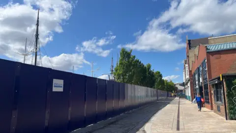 A large blue construction fence runs alongside a cobbled street obscuring the view of large trees and ships moored in Hull Marina. A woman, dressed in blue, is walking away from the camera. Several buildings are visible on the right of the image.