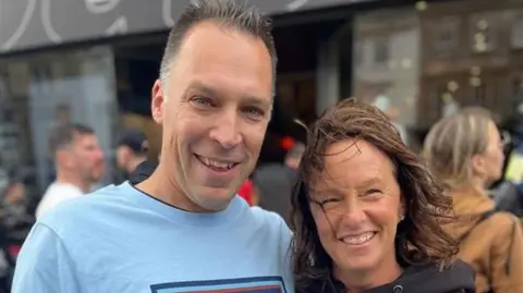 Teddy Cheesebrough on the left wearing a blue Oasis t-shirt. He is smiling at the camera and has short dark grey hair. Carly Cheesebrough on the right with hair blowing over her face. She is smiling and is wearing a black hooded sweatshirt.