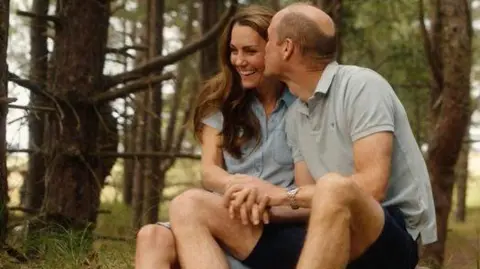 Catherine and William, the Princess and Prince of Wales, sit together holding hands in a forest setting for a colour photograph showing him kissing her as she smiles in an image released for Valentine's Day.