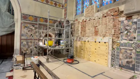 Jamie Niblock/BBC Wall bare of mosaic exposes brick and wood panelling. Two workmen stand on scaffolding. Other examples of mosaic wall-coverings can be seen on two walls in the altar area of the church. A large multi-coloured stained-glass window is above the chancel.