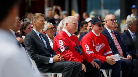 Oficiali canadieni - ministrul infrastructurii Gregor Robertson într-un costum gri, Doug Ford într-un tricou roșu cu o frunză de arțar neagră și primarul orașului Windsor, Drew Dilkens, într-o șapcă de baseball neagră și un tricou roșu al echipei Red Wings, stau la rând.